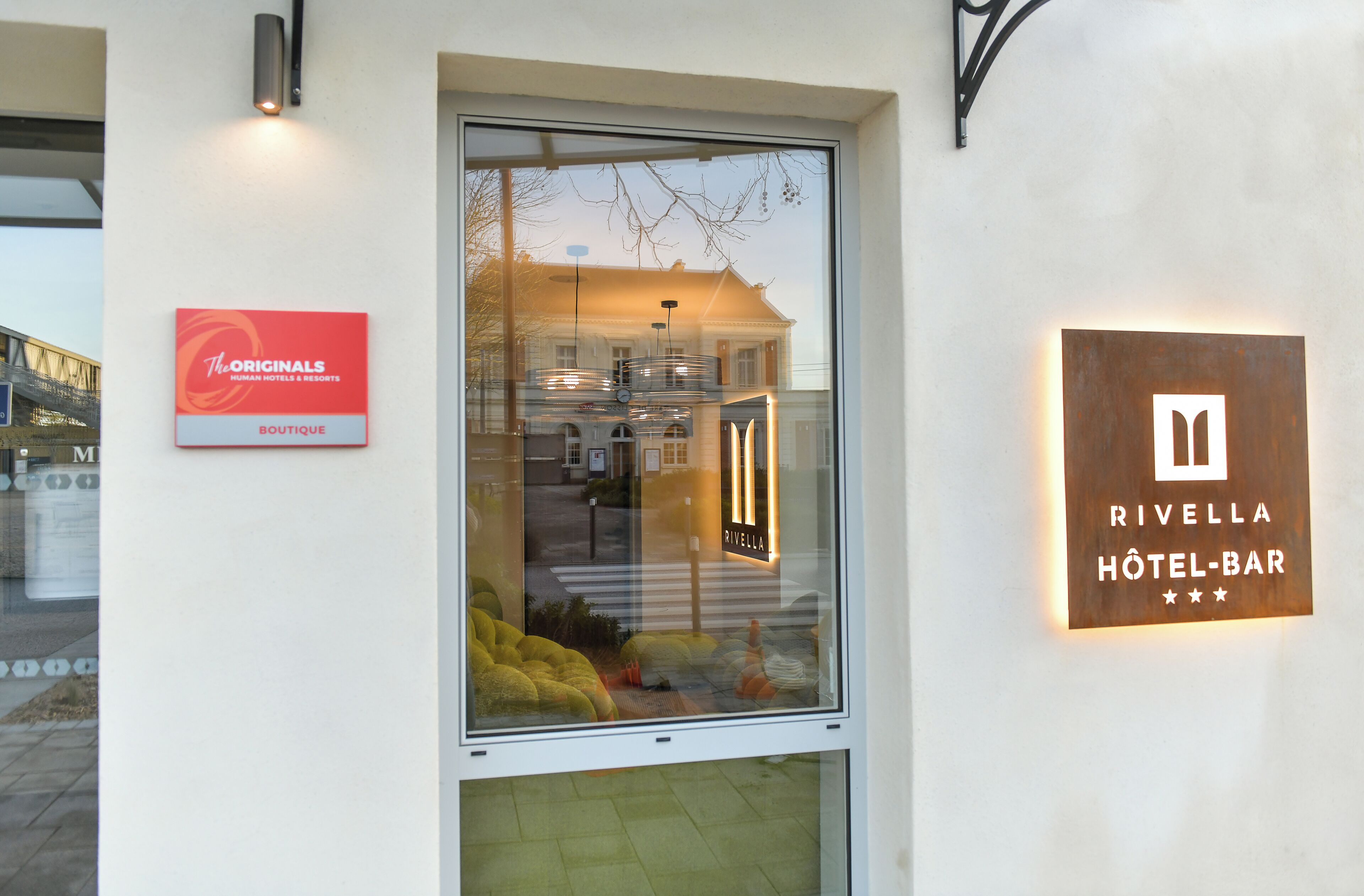 Entrada de un hotel con puerta de cristal, cartel de 'The Originals Boutique' y cartel de 'Rivella Hotel-Bar'.