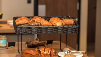 Une table en bois dressée pour le petit-déjeuner, avec des croissants, une miche de pain et une assiette sur laquelle se trouve une pâtisserie.