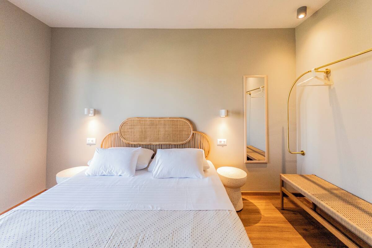 A bedroom with a large bed, bedside tables, a bench, a mirror, and a shower area.