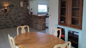 A dining area with a round wooden table, six chairs, a hanging light fixture, a stone wall, a kitchen in the background, and a wooden cabinet with glass doors. - Vacation home 3 km from the sea, in Brittany in Plestin-les-Grèves (plestin les grèves)