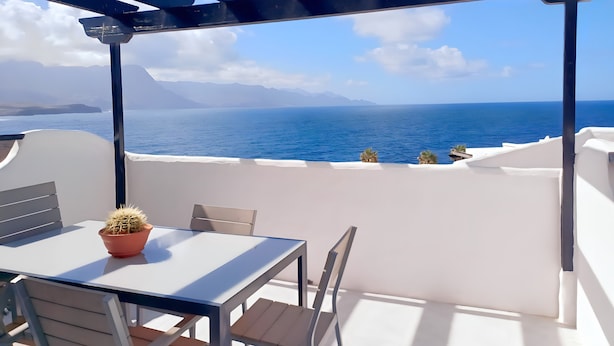 Una terraza con mesa y sillas blancas, una planta en maceta y vistas al mar y las montañas.