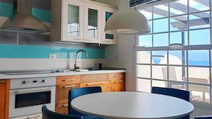 Una cocina moderna con una mesa redonda y sillas azules, un ventanal con vista al mar y un ventilador de techo blanco.