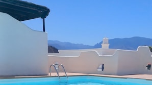 Piscina azul cristalina con escalera, rodeada de muros blancos y vistas a la montaña.