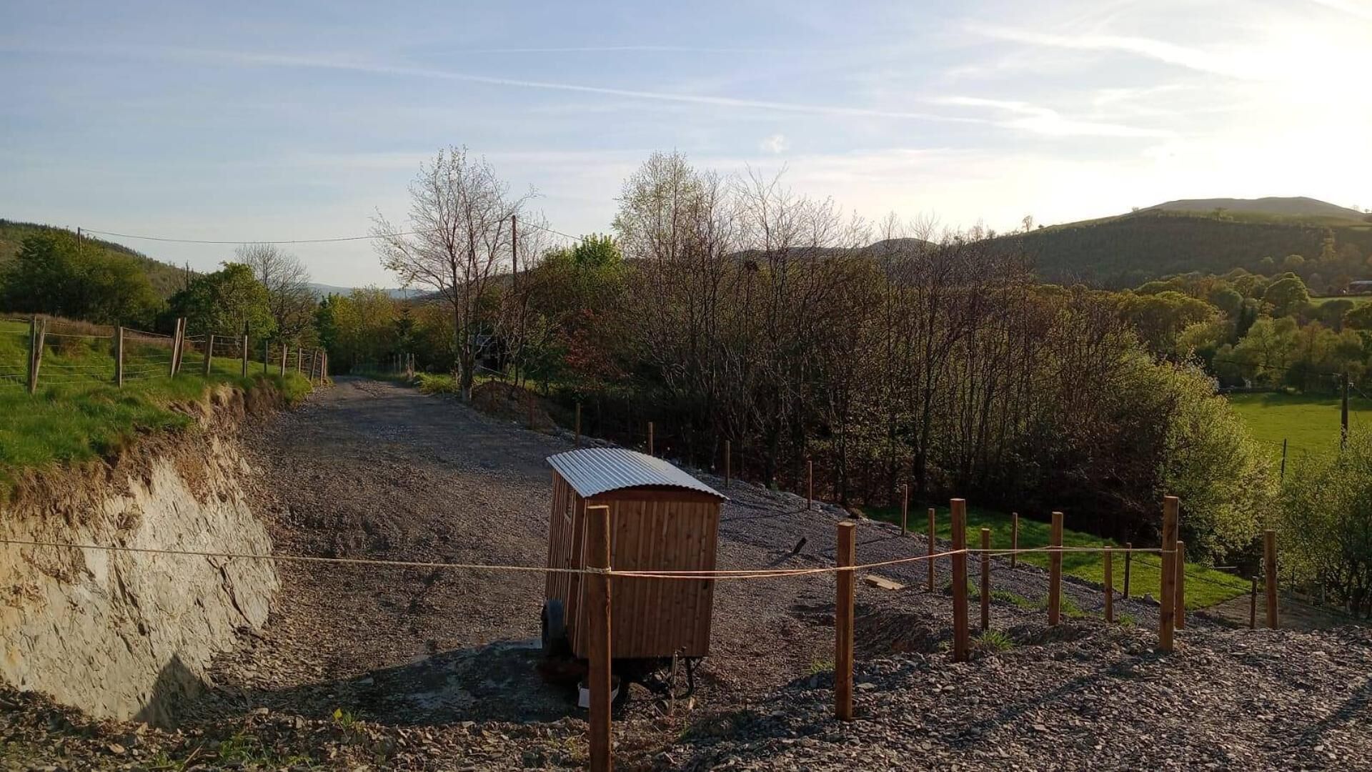 Un chemin de gravier mène à une structure en bois surmontée d’un toit en tôle ondulée, le tout étant entouré d’une clôture et d’arbres.