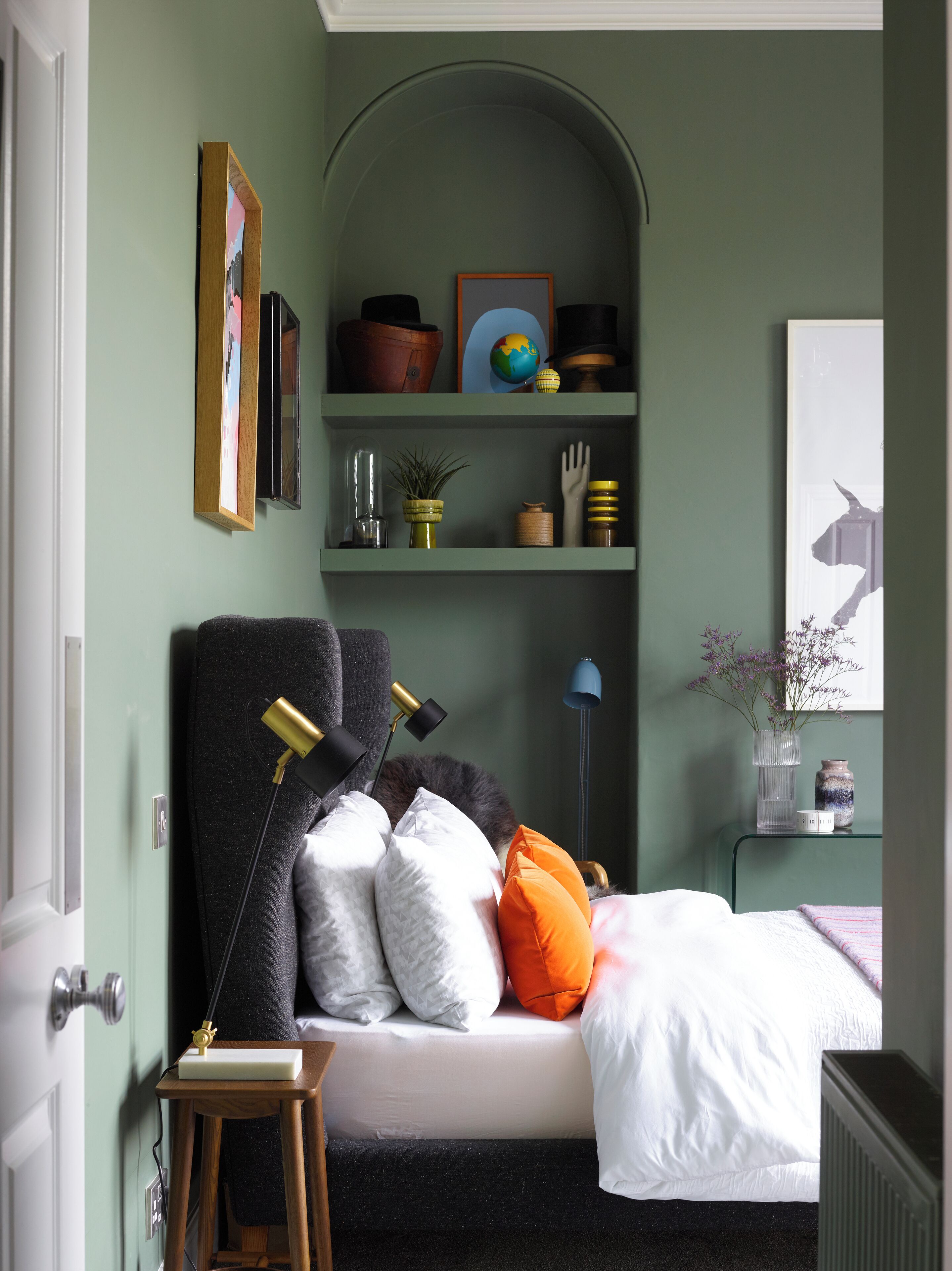 A bedroom with a bed, a bedside table, a lamp, a shelf with decorative items, and a door.