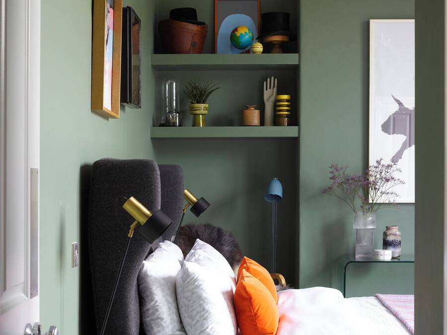 A bedroom with a bed, a bedside table, a lamp, a shelf with decorative items, and a door.