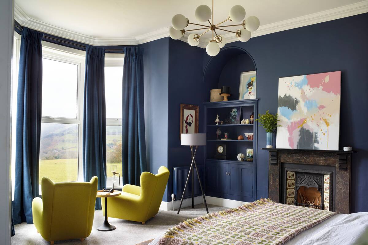Un dormitorio con una cama grande, un sillón amarillo, un sillón azul, una estantería y una chimenea.