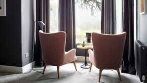 Ein Zimmer mit zwei Sesseln, einem kleinen Tisch und einem großen Fenster mit dunklen Vorhängen.
