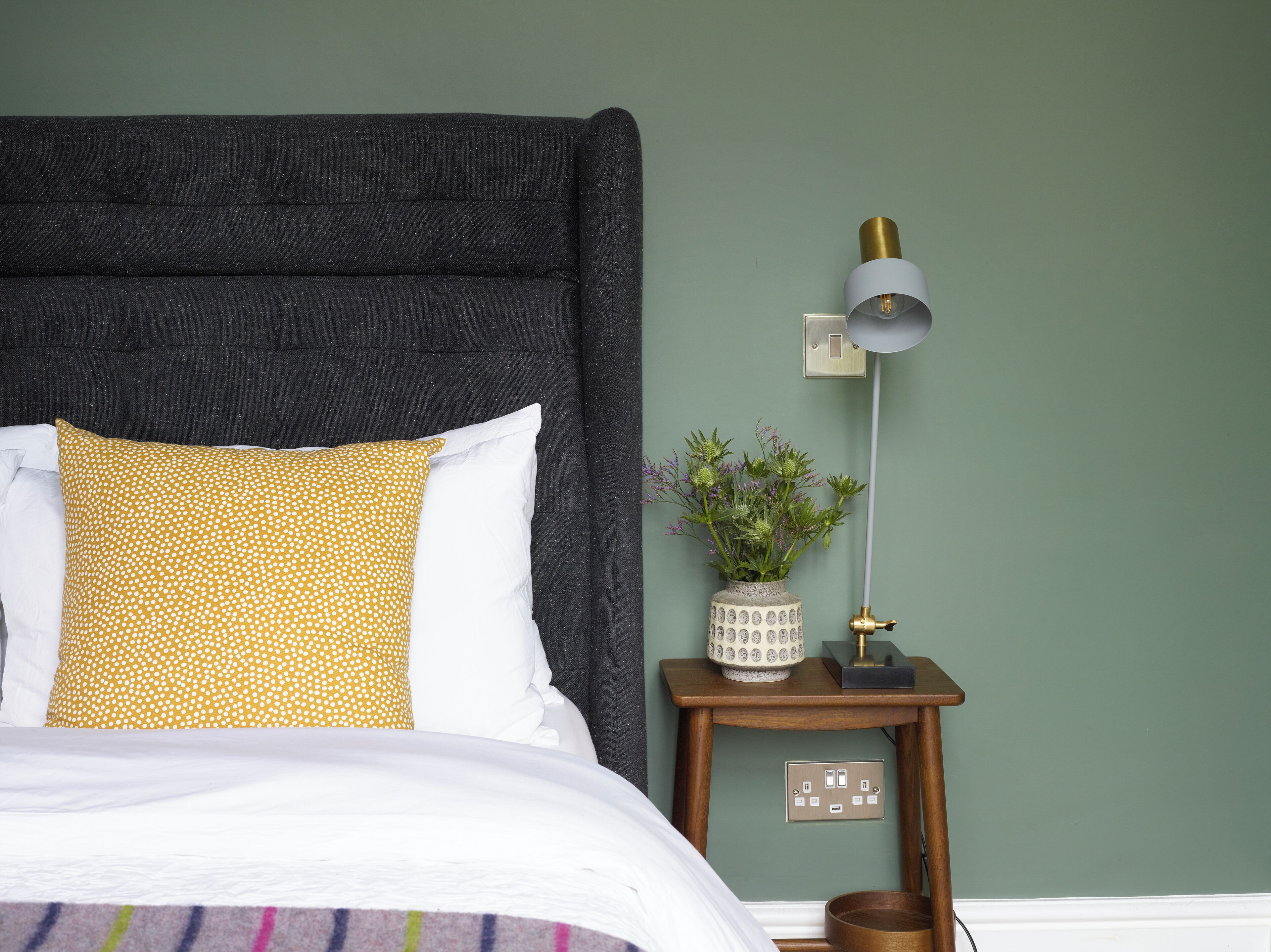 A bed with a dark headboard, white and yellow pillows, a small bedside table with a lamp, a vase with flowers, and a switchboard.