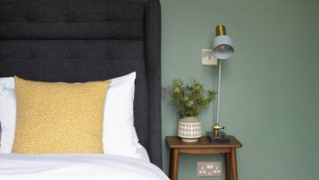 Un letto con testiera scura, cuscini bianchi e gialli, un comodino con una lampada, un vaso con fiori e un quadro elettrico.