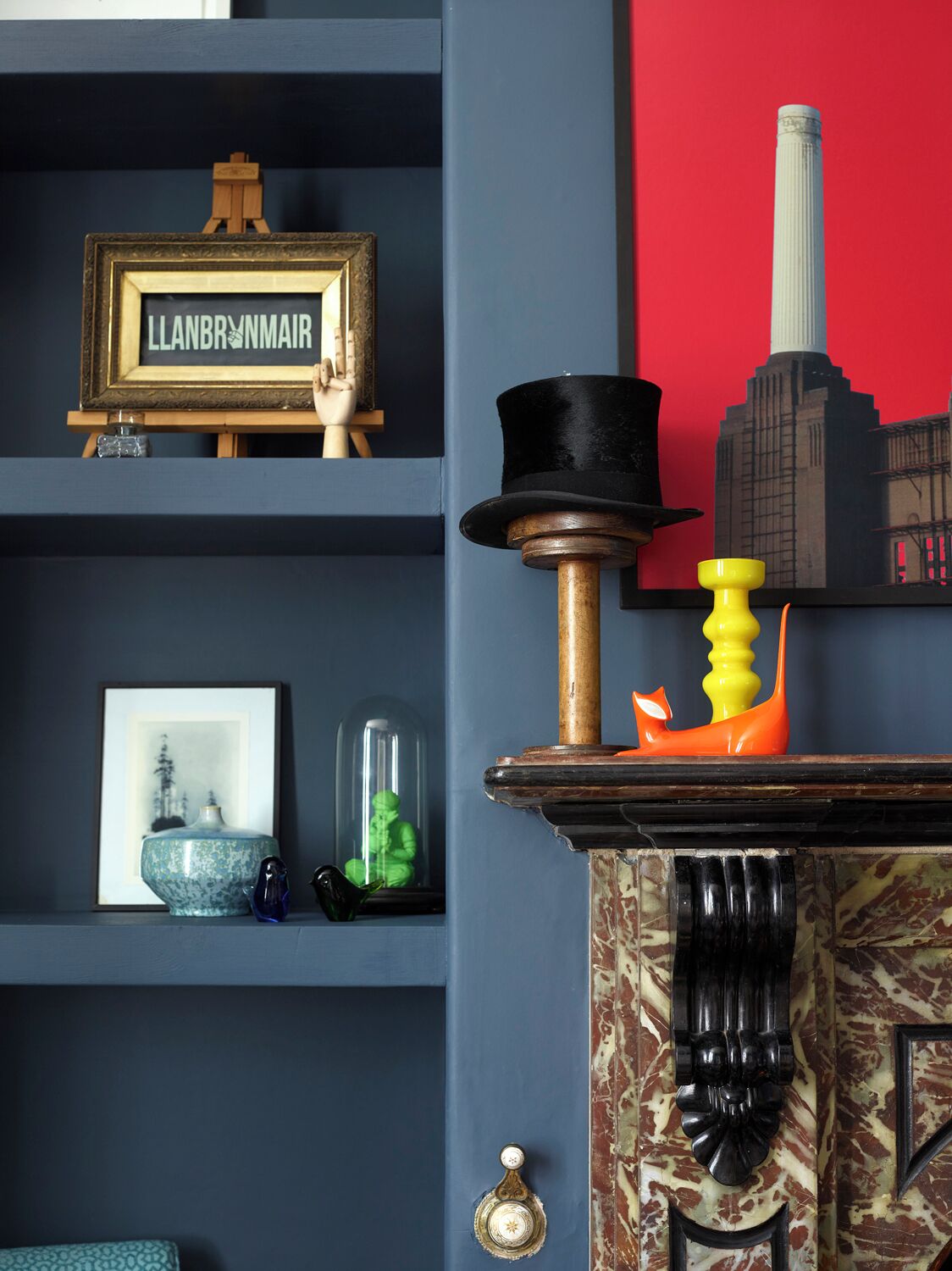 Une pièce avec un mur bleu, une plaque nominative encadrée, un petit chevalet en bois, un tableau encadré, un vase, une figurine verte, un globe en verre, un objet jaune, un objet noir et un mur rouge orné de la silhouette d’un bâtiment.