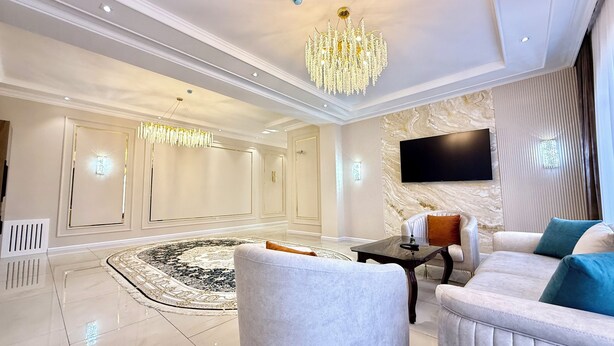 A spacious living room with a large chandelier, a flat-screen TV mounted on the wall, and a plush sofa with decorative pillows. - Maksima (Samarkand)