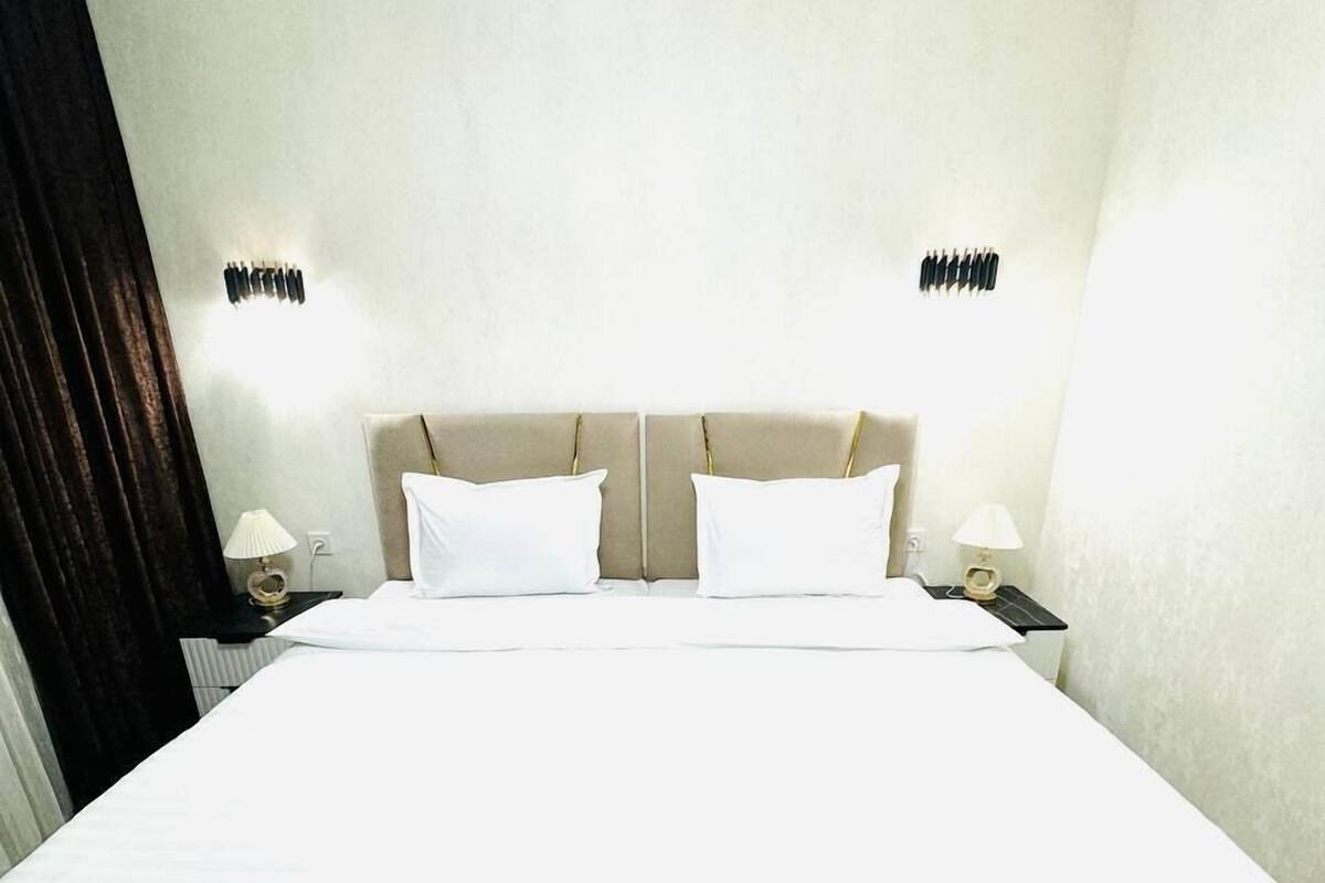 Una cama bien hecha con un cabecero color beige, dos mesitas de noche con lámparas y un aplique de pared.