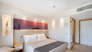 A modern hotel room with a large bed, a bedside table, a nightstand, a wall-mounted lamp, and a painting of a sunset.