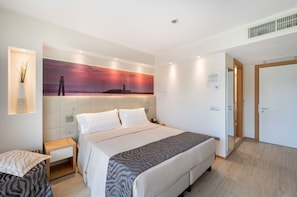 Une chambre d’hôtel moderne avec un grand lit, une table de chevet, une petite table d’appoint, une lampe fixée au mur et un tableau représentant un coucher de soleil.