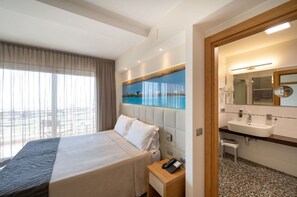 Habitación de hotel con cama, mesita de noche con teléfono, ventana grande con cortinas, baño con lavamanos y espejo, y un cuadro en la pared que muestra un cielo azul y agua.