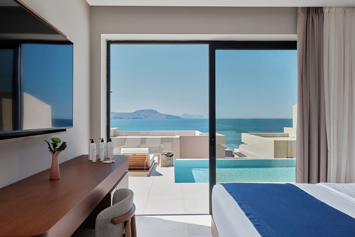 Una camera d'albergo con un letto, una scrivania con articoli da toilette, una sedia, vista sul mare e una piscina.