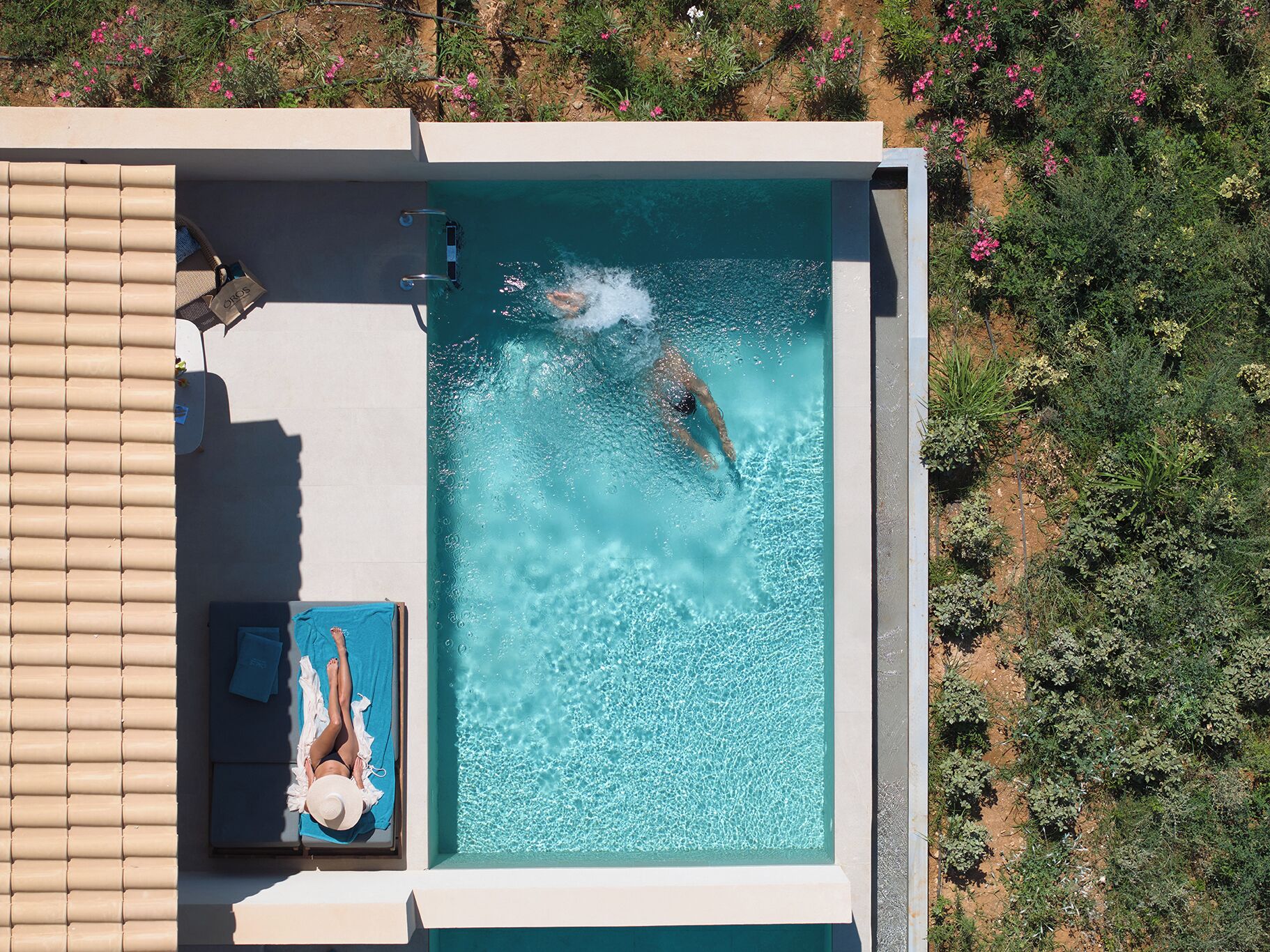 A rectangular swimming pool with a person swimming, a lounge area with a blue towel, and a view of surrounding vegetation.