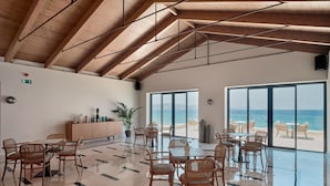 A spacious indoor area with a high ceiling, large windows offering a view of the sea, and a polished floor.