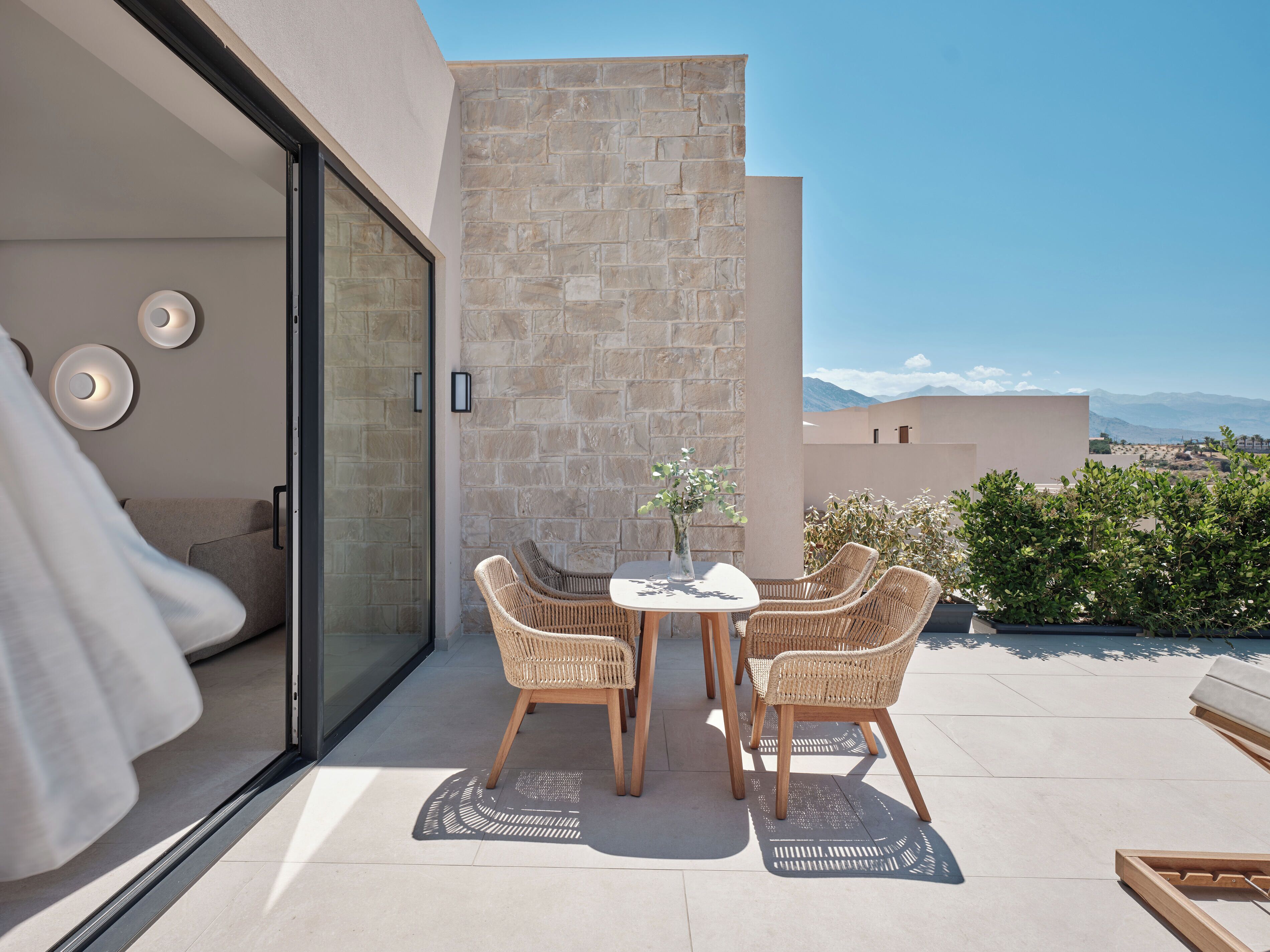 Un espace extérieur moderne, aménagé pour se détendre, avec une porte vitrée donnant sur un balcon, des murs en pierre et une vue sur les montagnes lointaines.