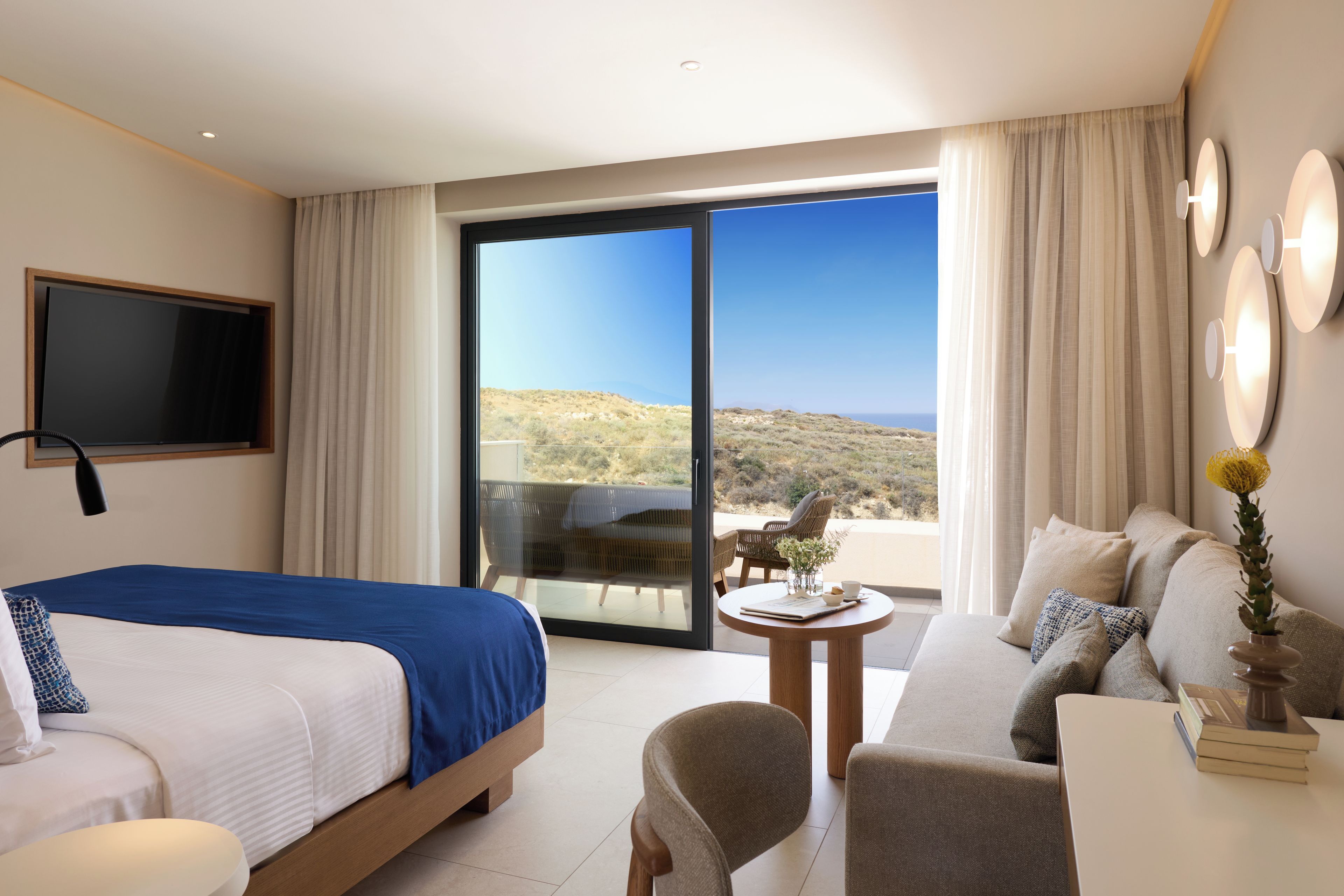 A hotel room with a bed, a sofa, a small table, and a balcony with a view of the landscape.