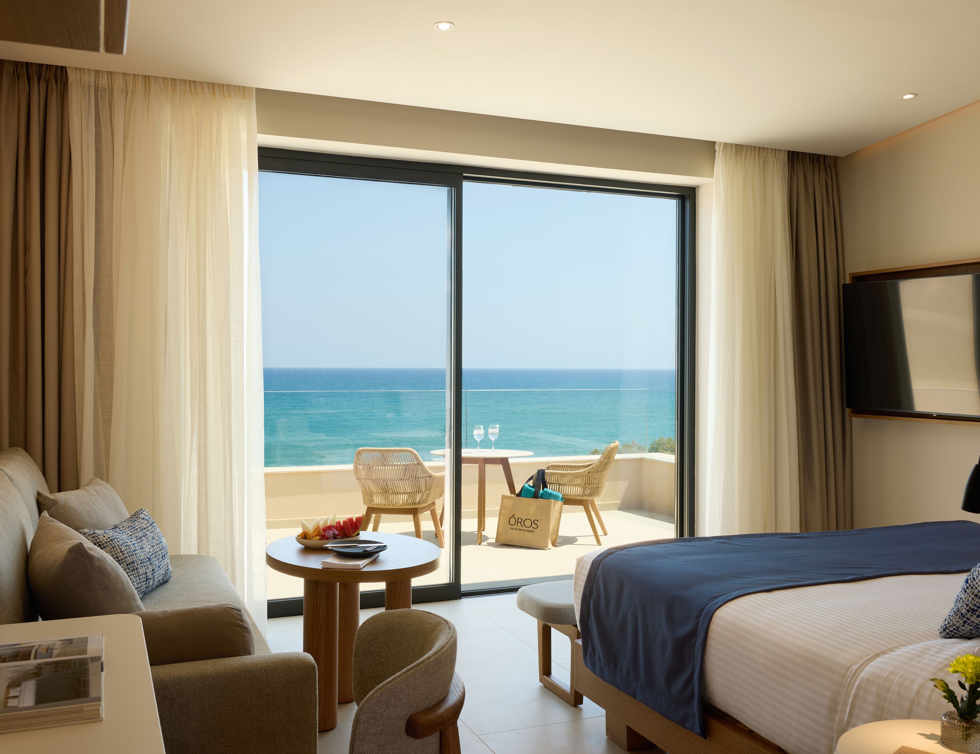 Habitación de hotel con cama, sofá, mesa de comedor con sillas y vista al océano a través de una puerta corrediza de vidrio.