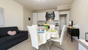 A modern living room with a black sofa, white chairs, and a dining table with a white lace tablecloth. - My Peschiera Holiday Apartment- Quadrivium 3 (Castelnuovo del Garda)