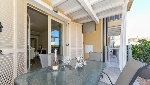 An outdoor dining area with a glass table, chairs, a bottle of wine, and a decorative sculpture. - My Peschiera Holiday Apartment- Quadrivium 3 (Castelnuovo del Garda)