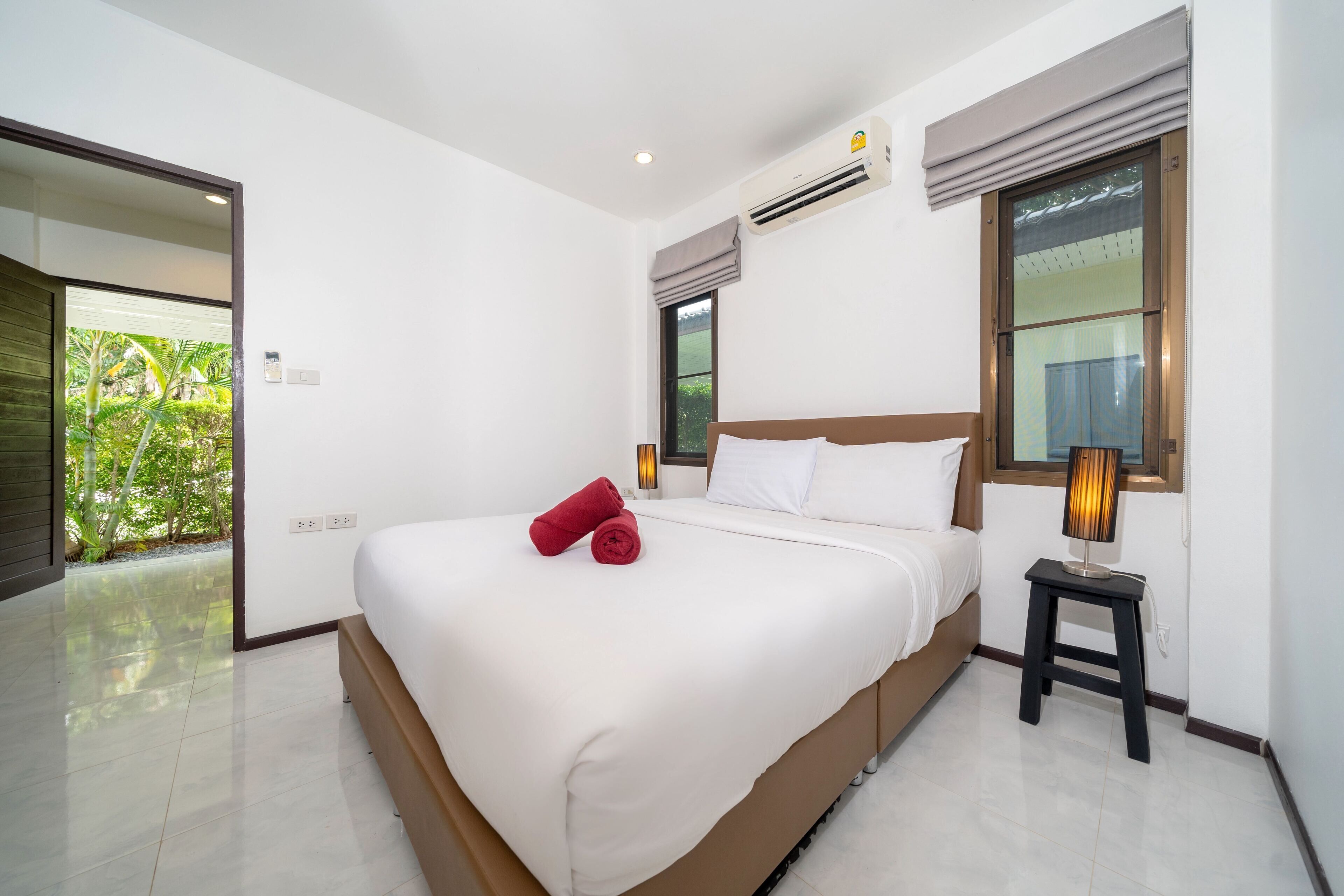 Habitación de hotel moderna con una cama grande, una mesita de noche pequeña con una lámpara, un equipo de aire acondicionado y una ventana con vistas a zonas verdes.