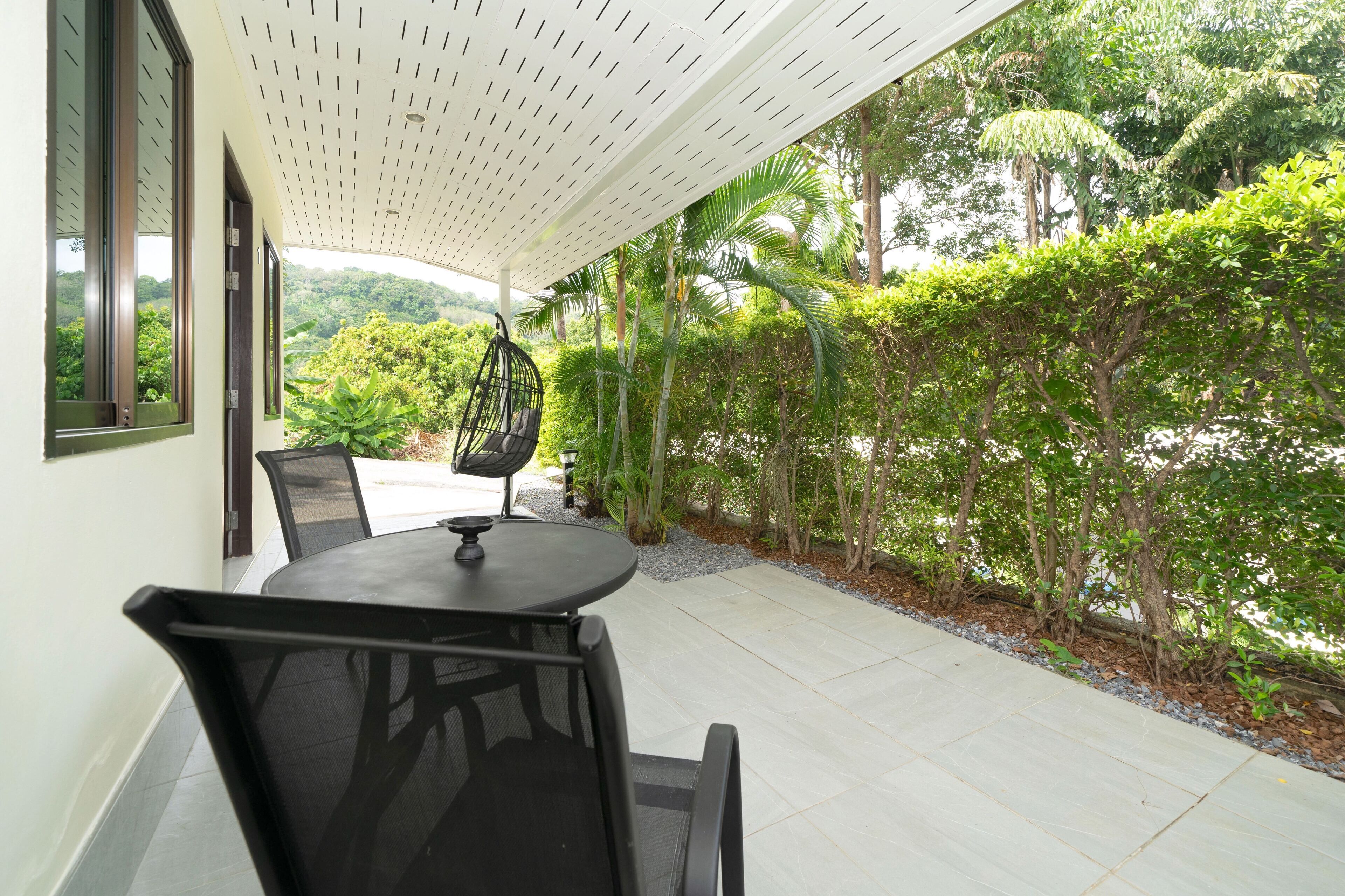Une terrasse extérieure avec une table ronde et des chaises, un fauteuil suspendu, et une vue sur une végétation luxuriante et des arbres.