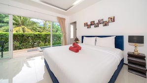 Una camera d'albergo moderna con un grande letto, un comodino con una lampada e vista su un verde rigoglioso attraverso ampie finestre.