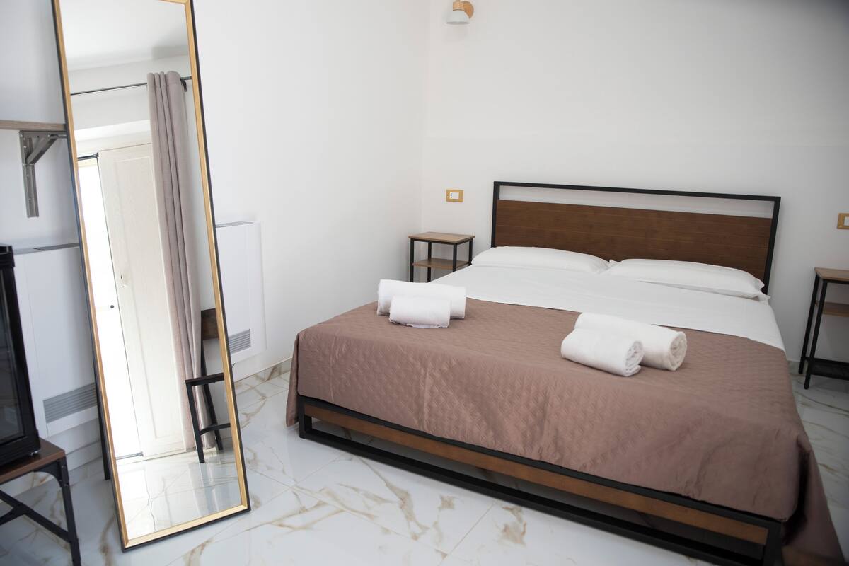 A neatly made bed with white and brown linens, a small bedside table, and a large mirror on the left.
