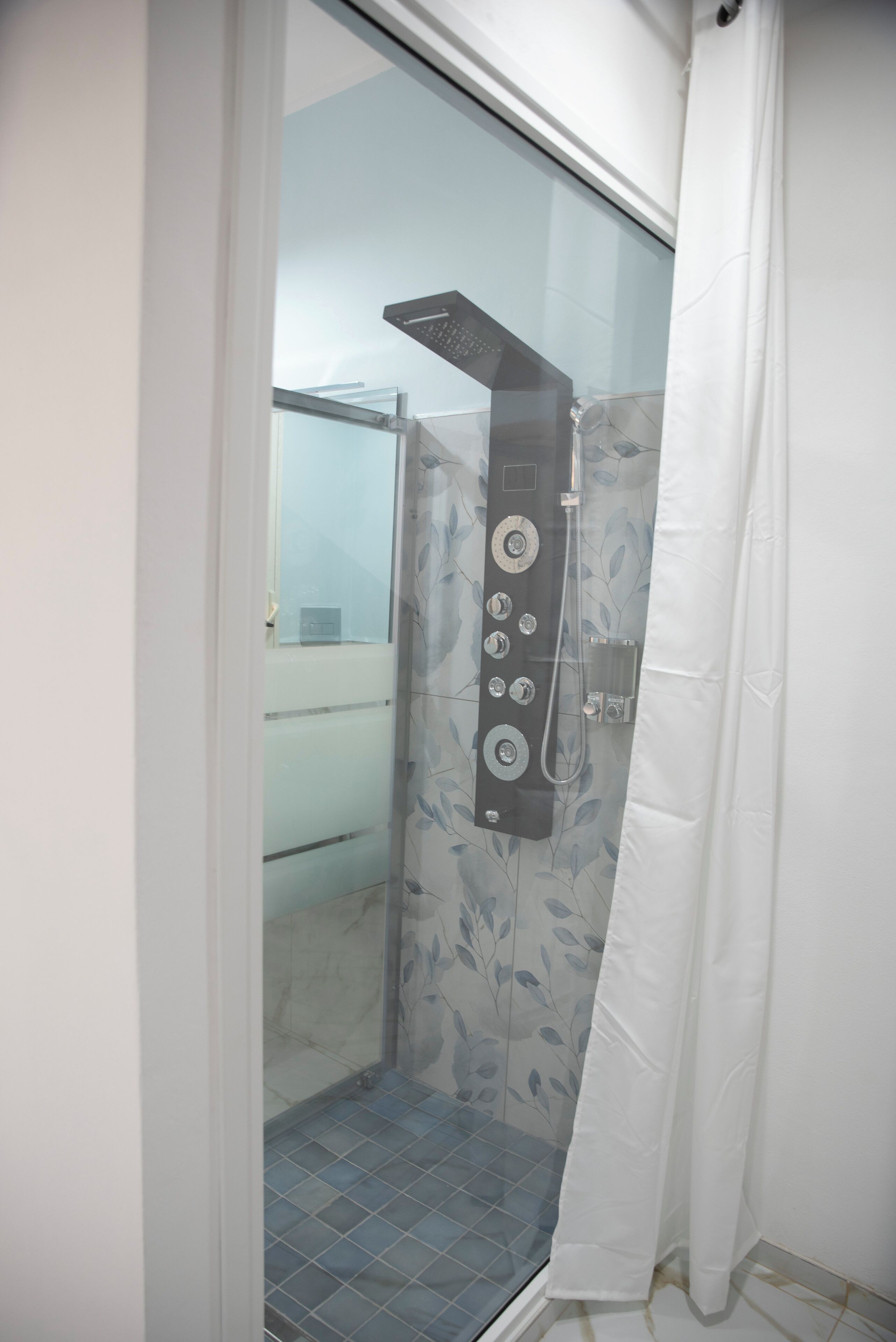 A glass-enclosed shower with a control panel, a white shower curtain, and a tiled floor.
