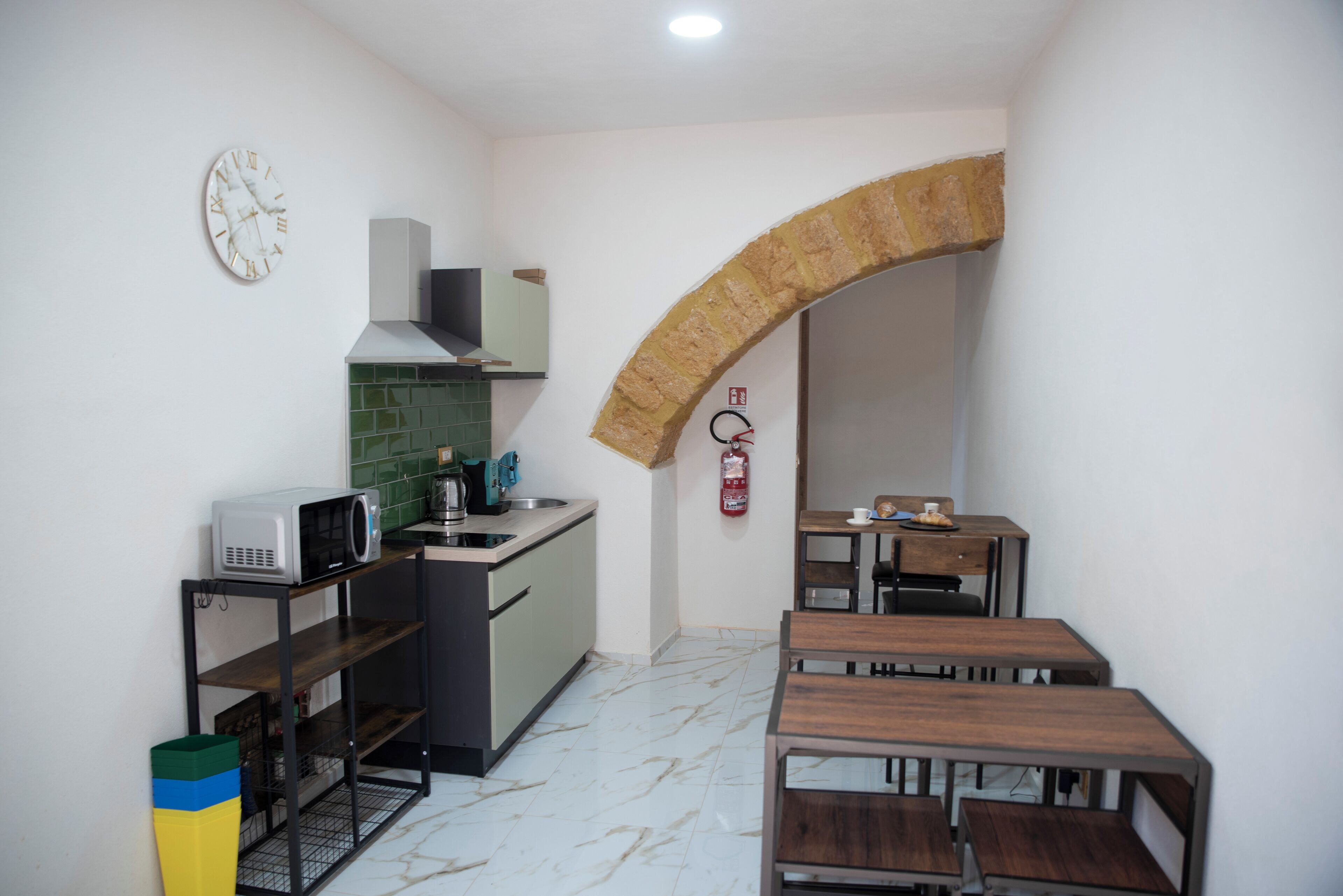 A compact kitchen with a microwave, sink, and green tiled backsplash. There's a wooden bench and a small table with a cup.