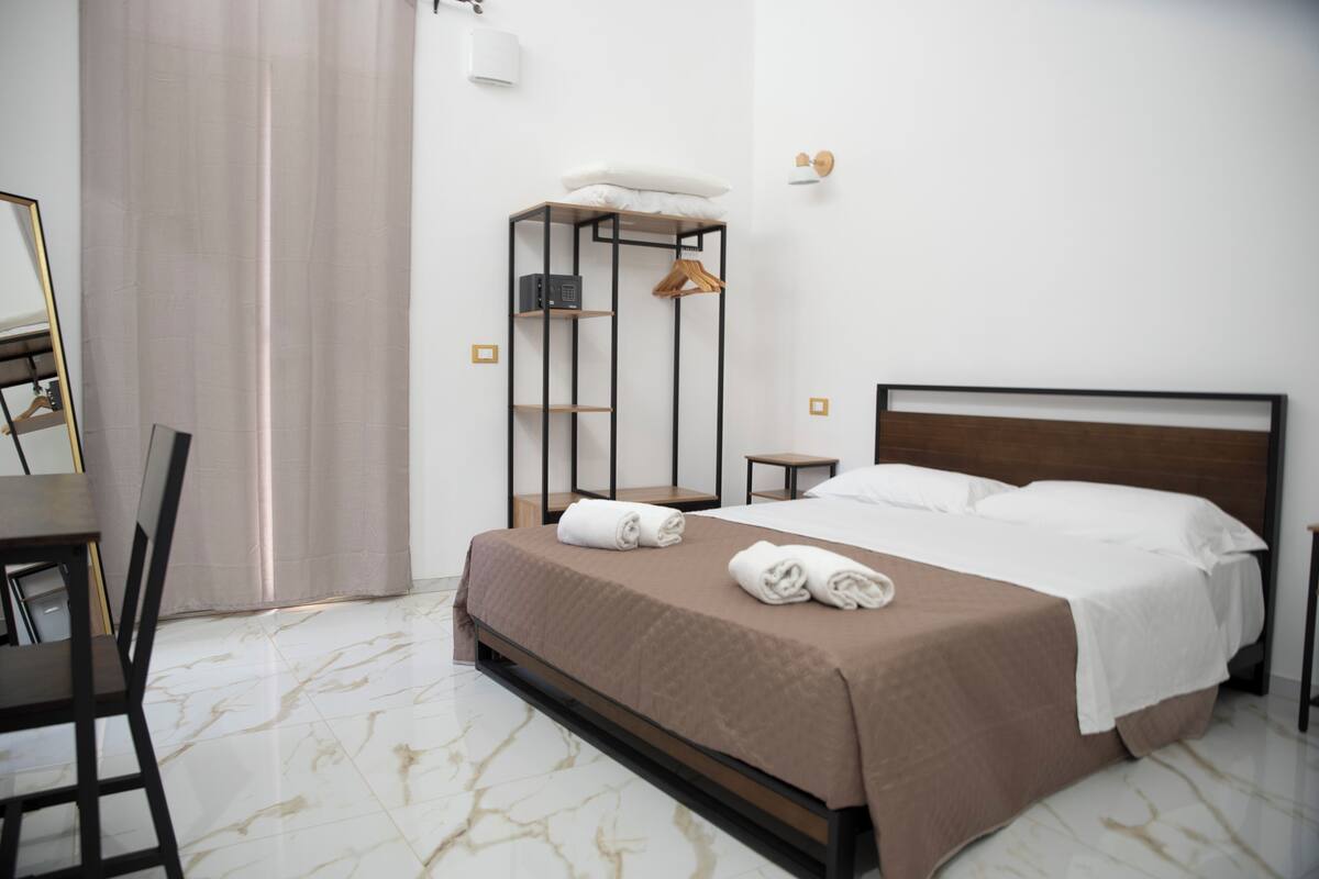 A neatly made bed with a brown bedspread and white linens, a bedside table with a lamp, a wall-mounted shelf with a hanging garment, and a chair.