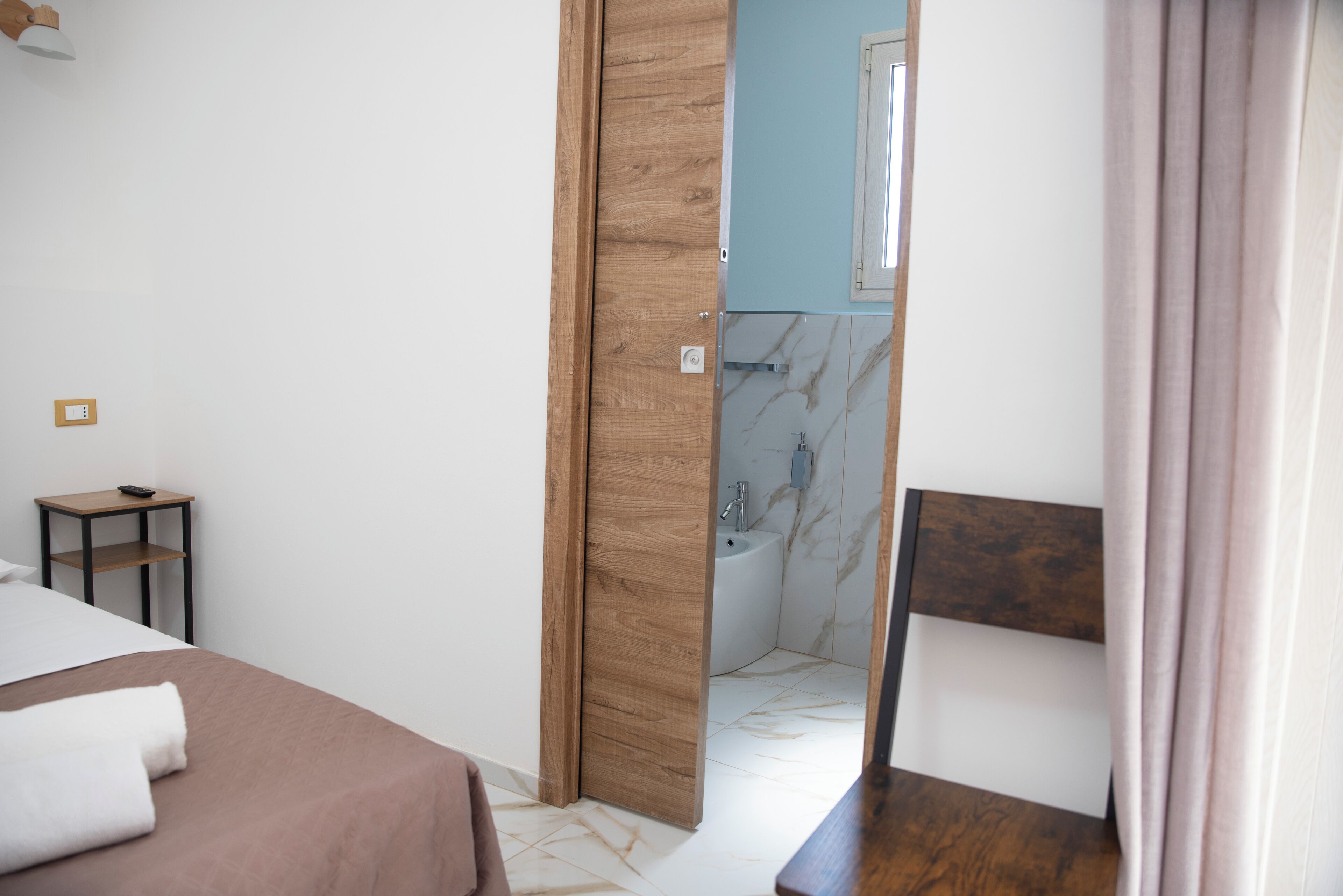 Quarto de hotel com uma cama, uma pequena mesa, uma porta de madeira que dá para um banheiro e um espelho.