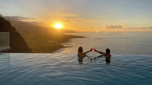 A couple toasting with wine glasses in a clear pool with a sunset view.