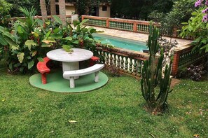 Mesa redonda con bancos en un jardín que tiene una piscina al fondo.