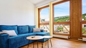 A room with a blue sofa, a small round table, and large windows offering a view of buildings and greenery. - Beautiful apartment for 4 people with WIFI and TV (Jilemnice)
