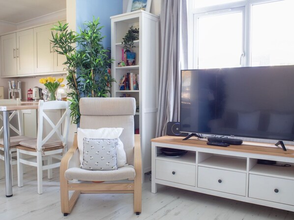 Una sala de estar moderna con un televisor de pantalla plana, un sillón cómodo, una estantería y un comedor con mesa y sillas.