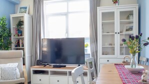 A living room with a TV on a wooden stand, a dining table with chairs, a cabinet with glass doors, and a plant. - Stunning private villa for 6 people with WIFI and TV (Isle Of Skye)