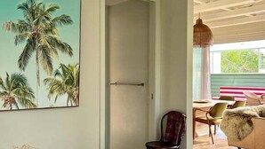 Un dormitorio con cama, un cuadro de palmeras, una puerta y vista hacia un comedor con sillas y una mesa.