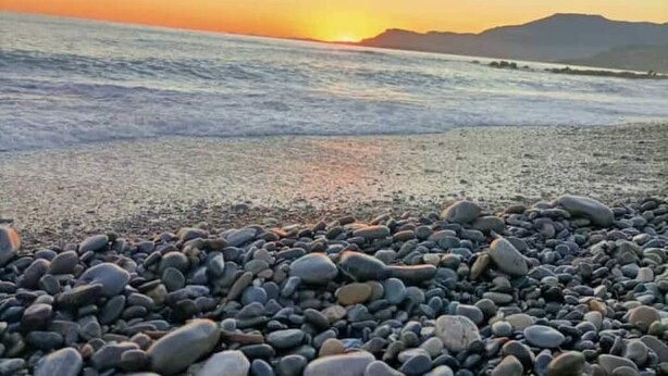 A pebbled beach with a sunset over the ocean and distant mountains.