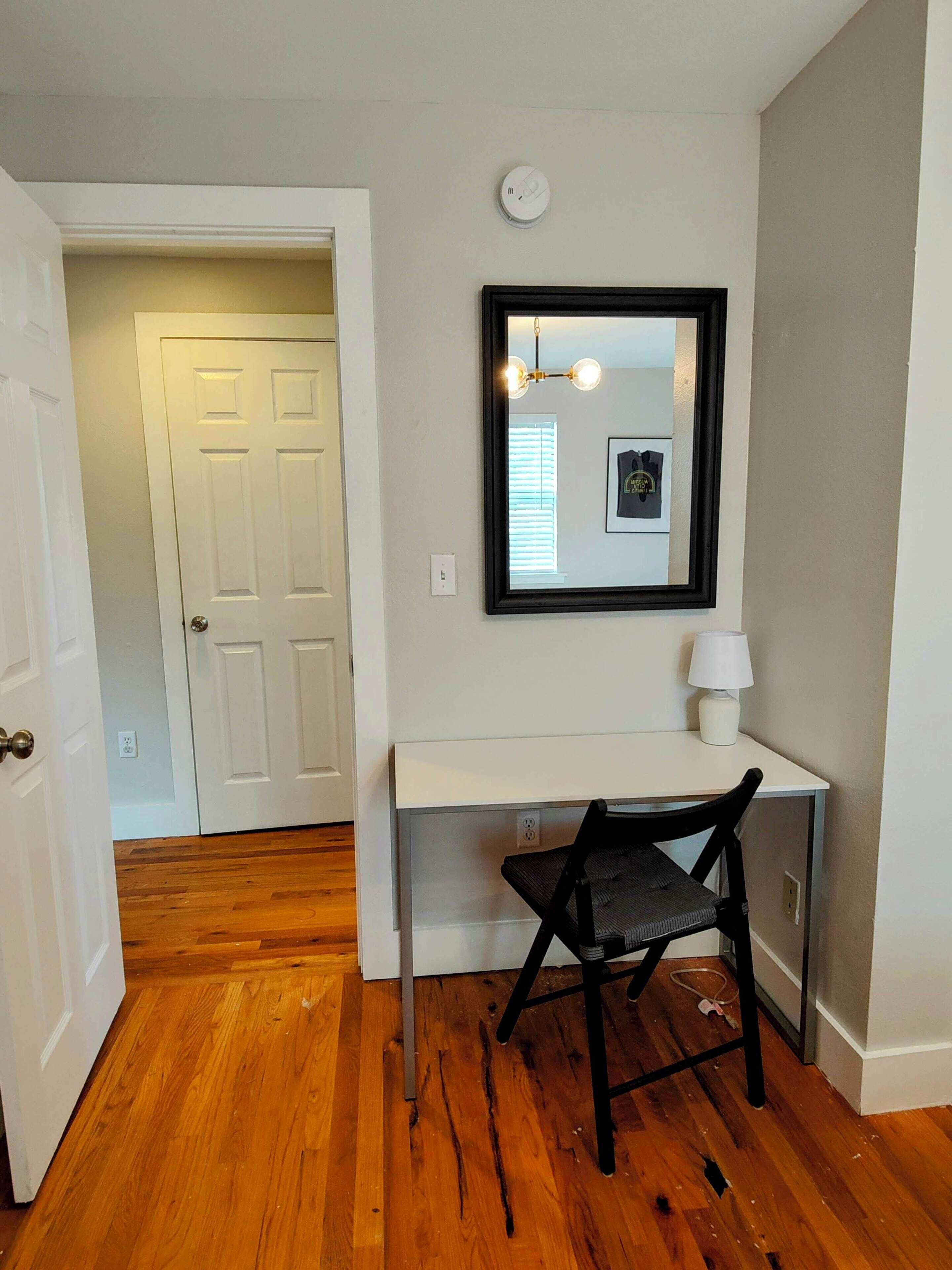 Une pièce avec un bureau blanc, une chaise noire, une lampe blanche et un tableau encadré accroché au mur.