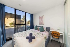 A hotel room with a bed, a desk, a chair, a window with curtains, and a wall-mounted picture.