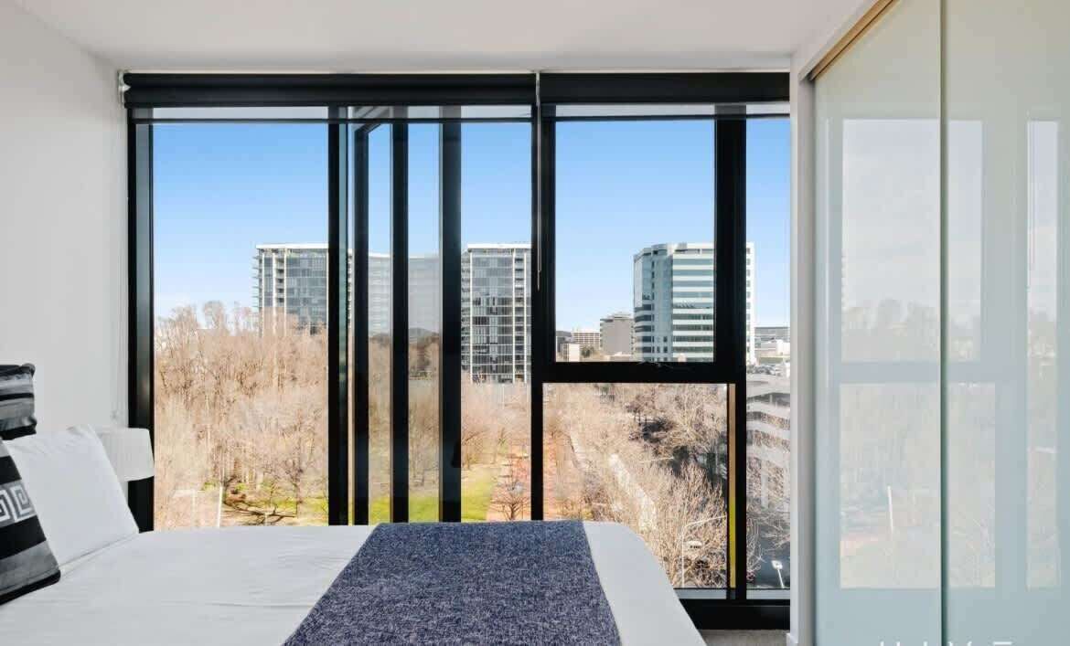 Une chambre avec une grande fenêtre, un lit et une vue sur des immeubles et des arbres.