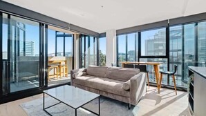 Ein modernes Wohnzimmer mit einer Couch, einem Couchtisch und einem Essbereich mit Tisch und Stühlen. Große Fenster bieten einen Blick auf die Stadt.