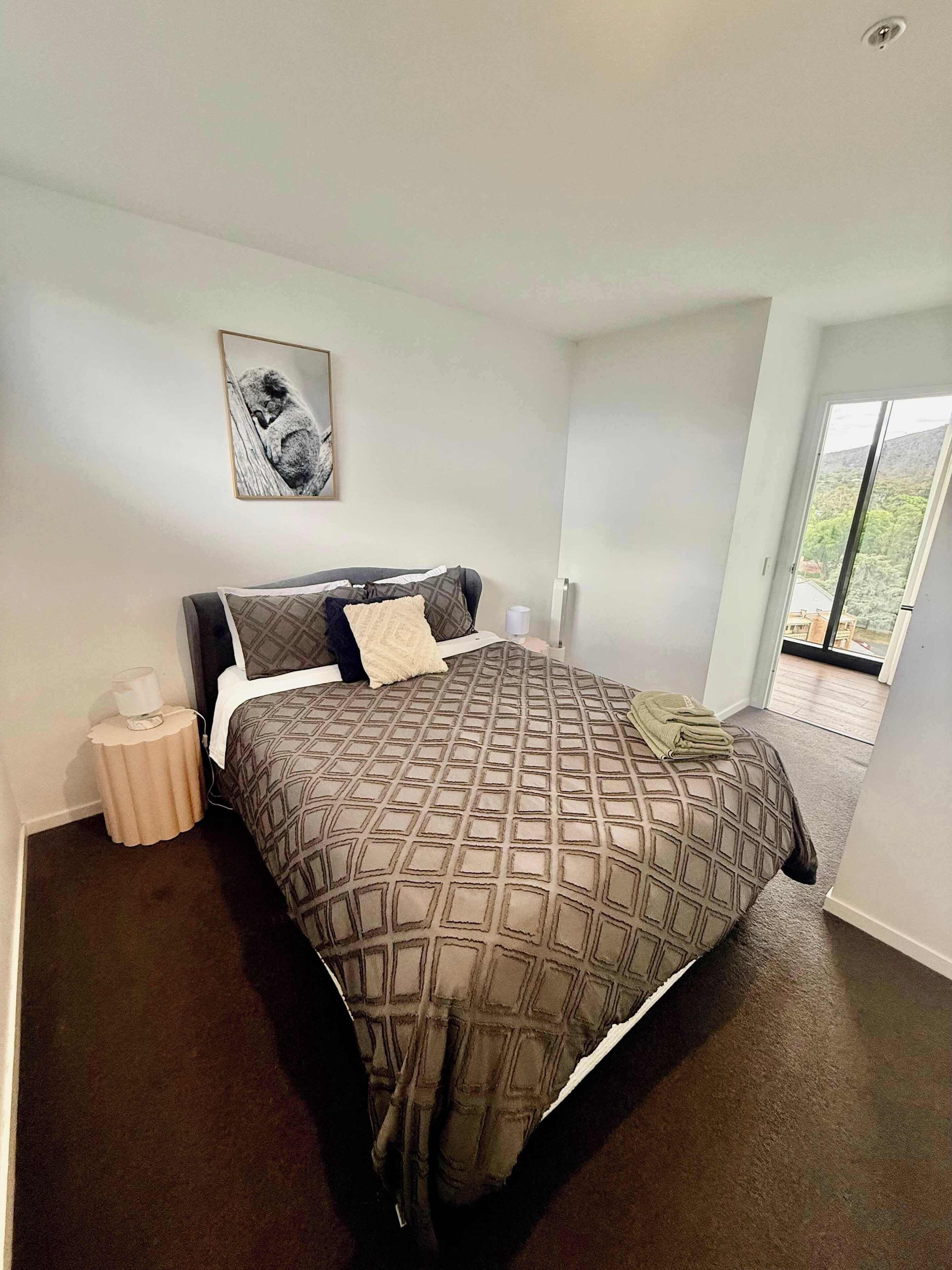 A bedroom with a large bed, a bedside table with a lamp, a framed picture on the wall, and a view of the outdoors through a sliding door.