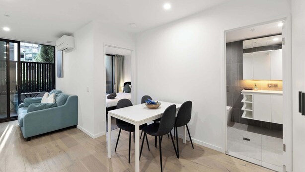 Une chambre d’hôtel moderne avec un coin repas, un canapé et une cuisine visible à travers une porte ouverte.