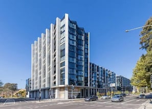 A modern multi-story building with a glass facade, located at a street corner with cars parked in front.